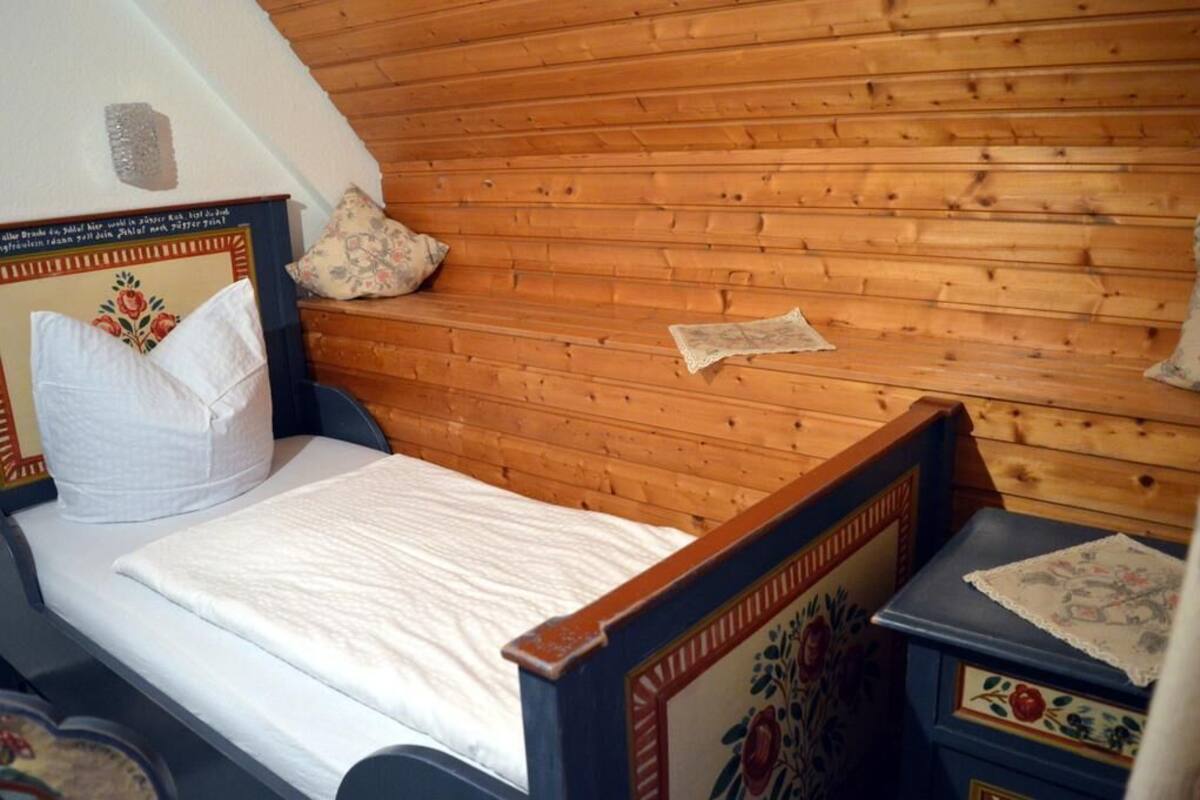 Een slaapkamer met een houten schuine plafond, een bed met een bloemen hoofdbord en een klein nachtkastje met een decoratieve doos.