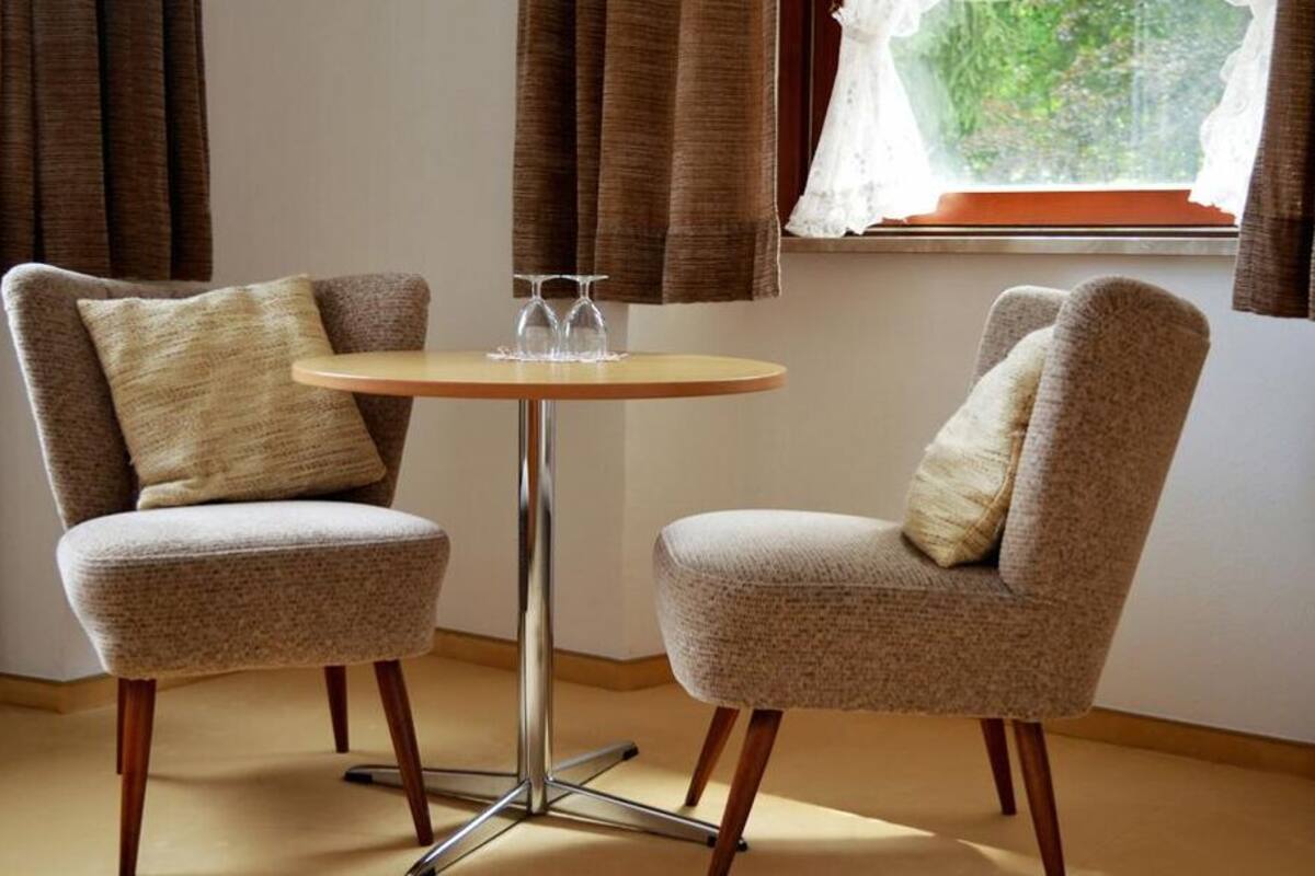 Twee fauteuils met een ronde tafel ertussen, met een raam met gordijnen en uitzicht op groen buiten.