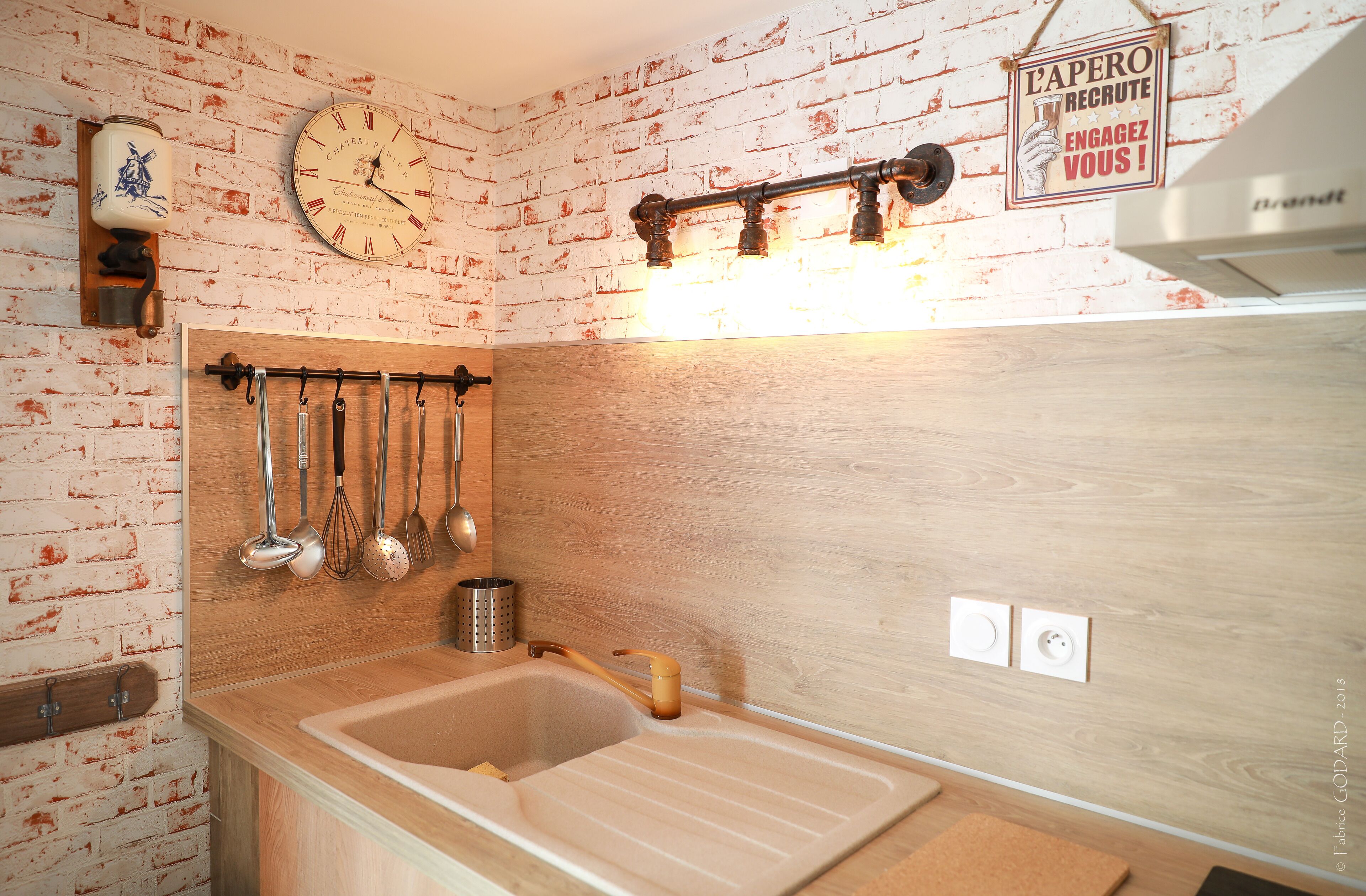 Une cuisine avec un plan de travail en bois, un étendoir suspendu où sont rangés les ustensiles, une horloge et un panneau accroché au mur.