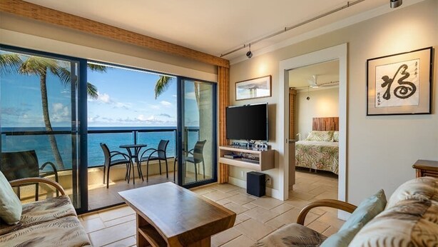 Ein modernes Wohnzimmer mit einer großen Schiebetür, die auf einen Balkon mit Meerblick führt. An der Wand ist ein Flachbildfernseher angebracht, es gibt einen Couchtisch aus Holz und ein Sofa mit gemusterten Kissen.