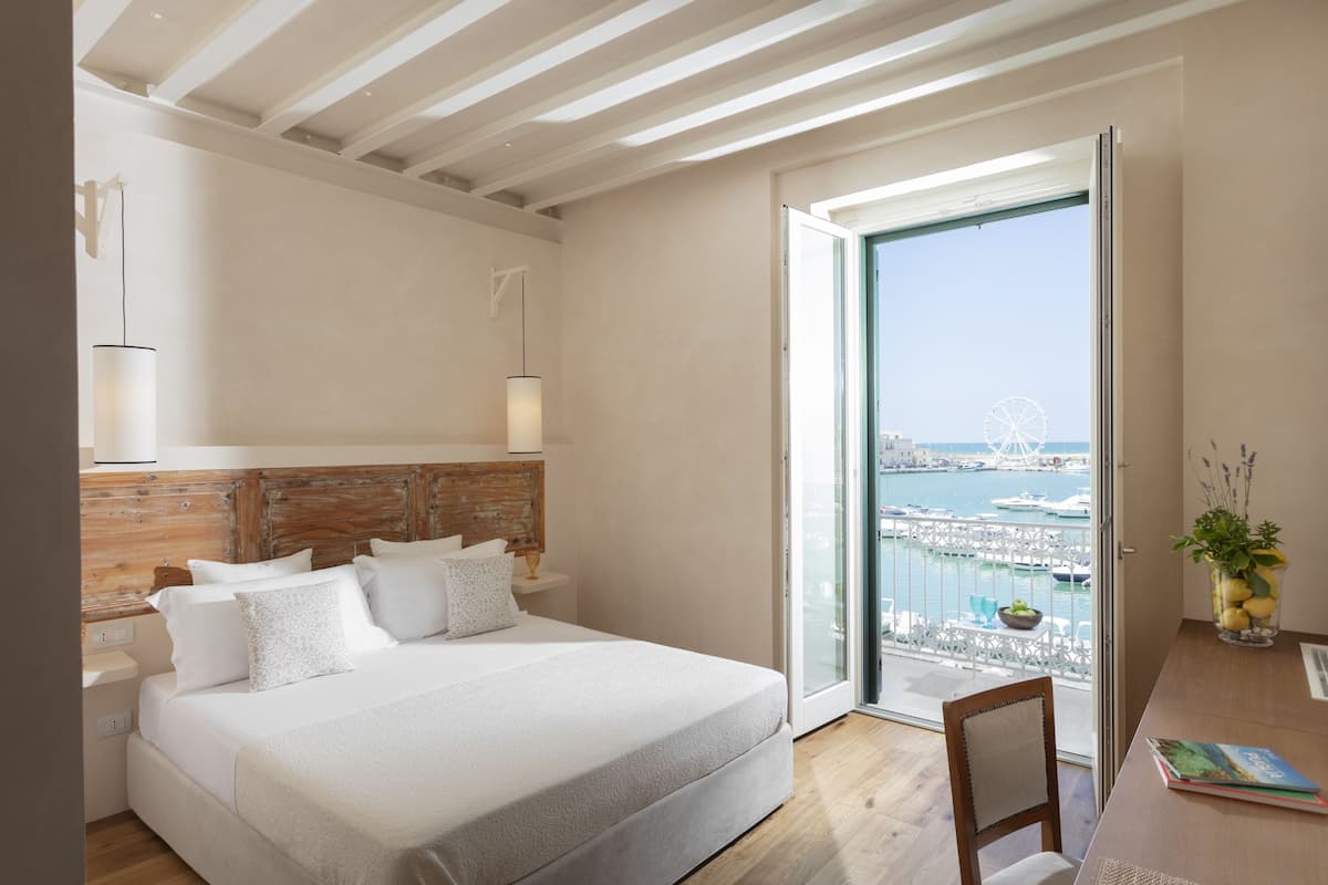 Un dormitorio con una cama grande, cabecero de madera, lámparas de noche, vista al puerto con barcos y un balcón con vista al puerto.