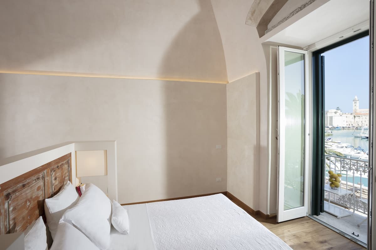 Habitación de hotel con una cama grande, cabecera de madera, mesita de noche y vista a la marina a través de una puerta de vidrio.