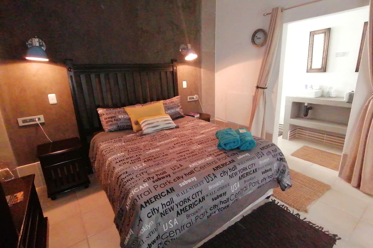 Habitación de hotel con cama, mesita de noche, lámpara, reloj y baño visible a través de una puerta abierta.
