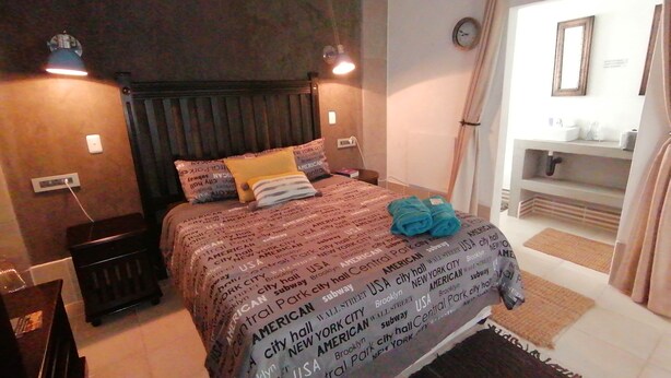 Habitación de hotel con cama, mesita de noche, lámpara, reloj y baño visible a través de una puerta abierta.