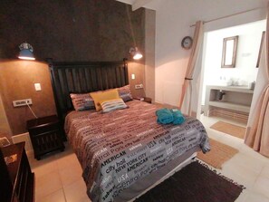 A hotel room with a bed, a nightstand, a lamp, a clock, and a bathroom visible through an open door.