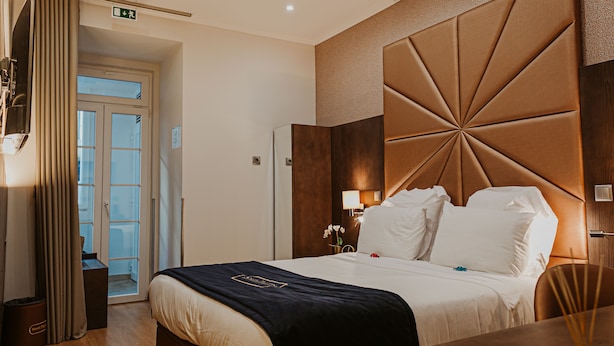 A hotel room with a large bed, a wooden headboard, a nightstand with a lamp, and a view of the outside through a door. - Seven Dreams Suites (Lisbon)