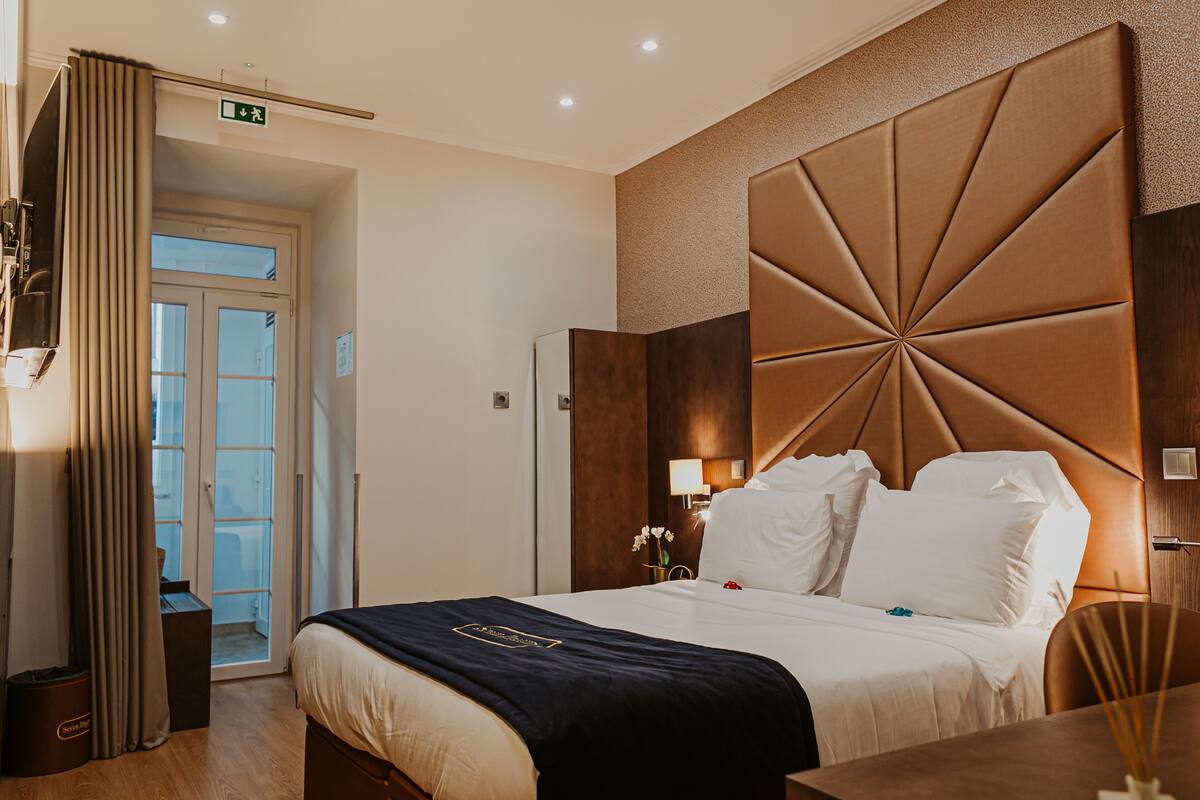 A hotel room with a large bed, a wooden headboard, a nightstand with a lamp, and a view of the outside through a door.