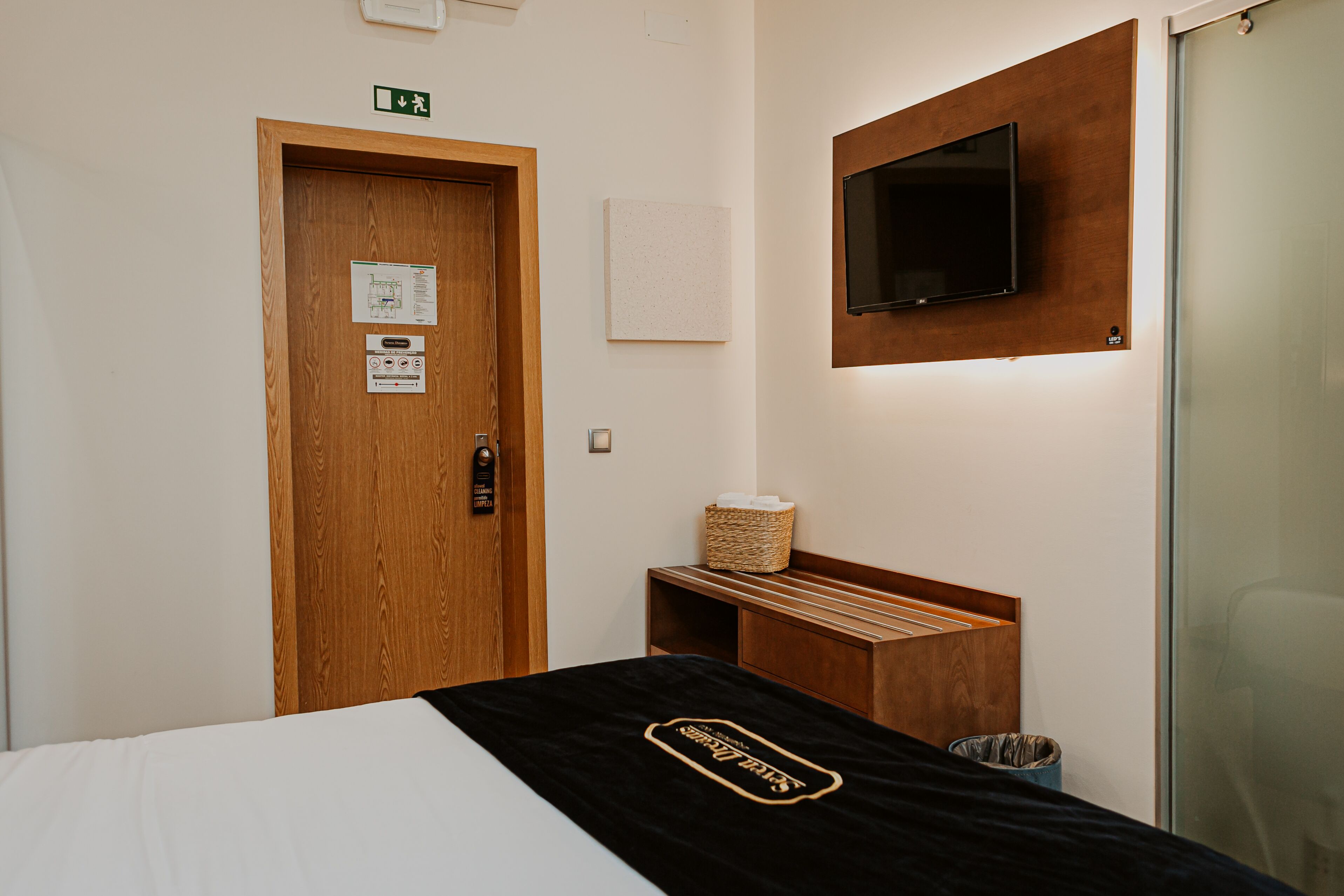 Habitación de hotel con puerta de madera, un televisor de pantalla plana montado en la pared, una cama con colcha negra, un pequeño banco de madera y un cesto de basura.