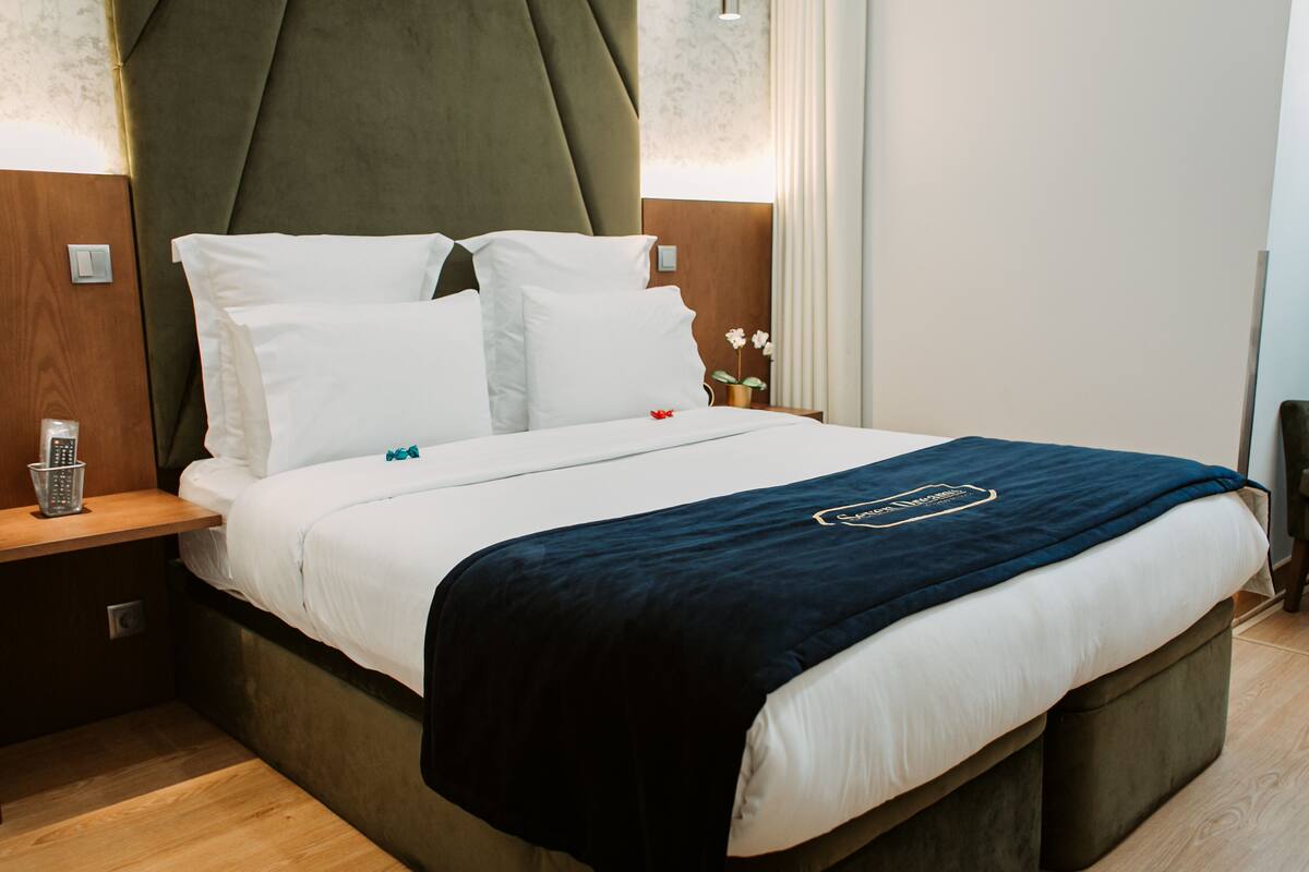 A neatly made bed with white linens and a dark blue blanket, a wooden nightstand with a glass of water and a remote control, and a green upholstered headboard.