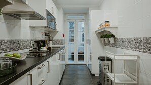 A narrow kitchen with white cabinets, a black countertop, and a tiled backsplash. There are shelves with jars and a trash bin. - Seven Dreams Suites (Lisbon)