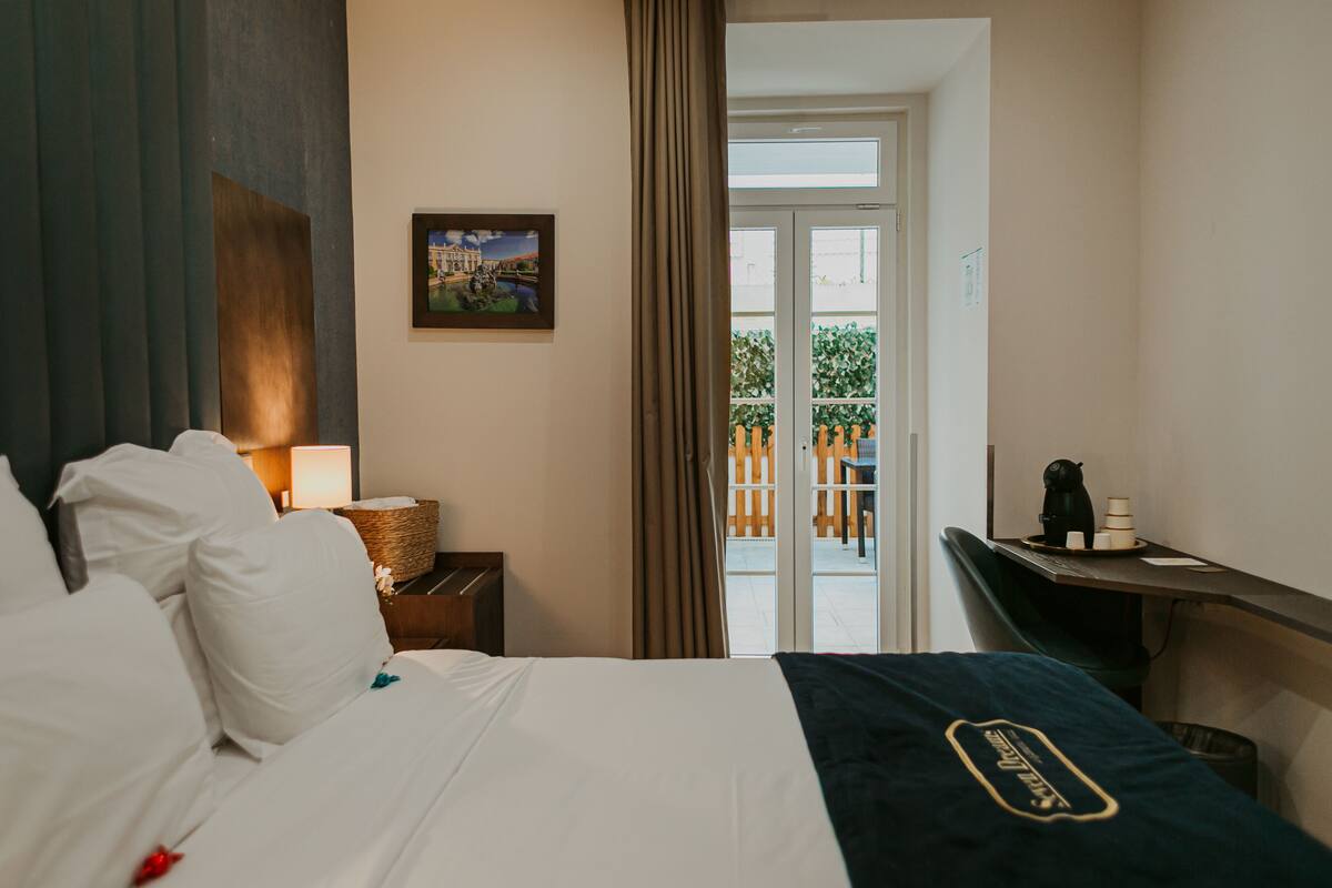 A hotel room with a bed, a nightstand with a lamp, a desk with a coffee maker, and a view of the outside balcony.