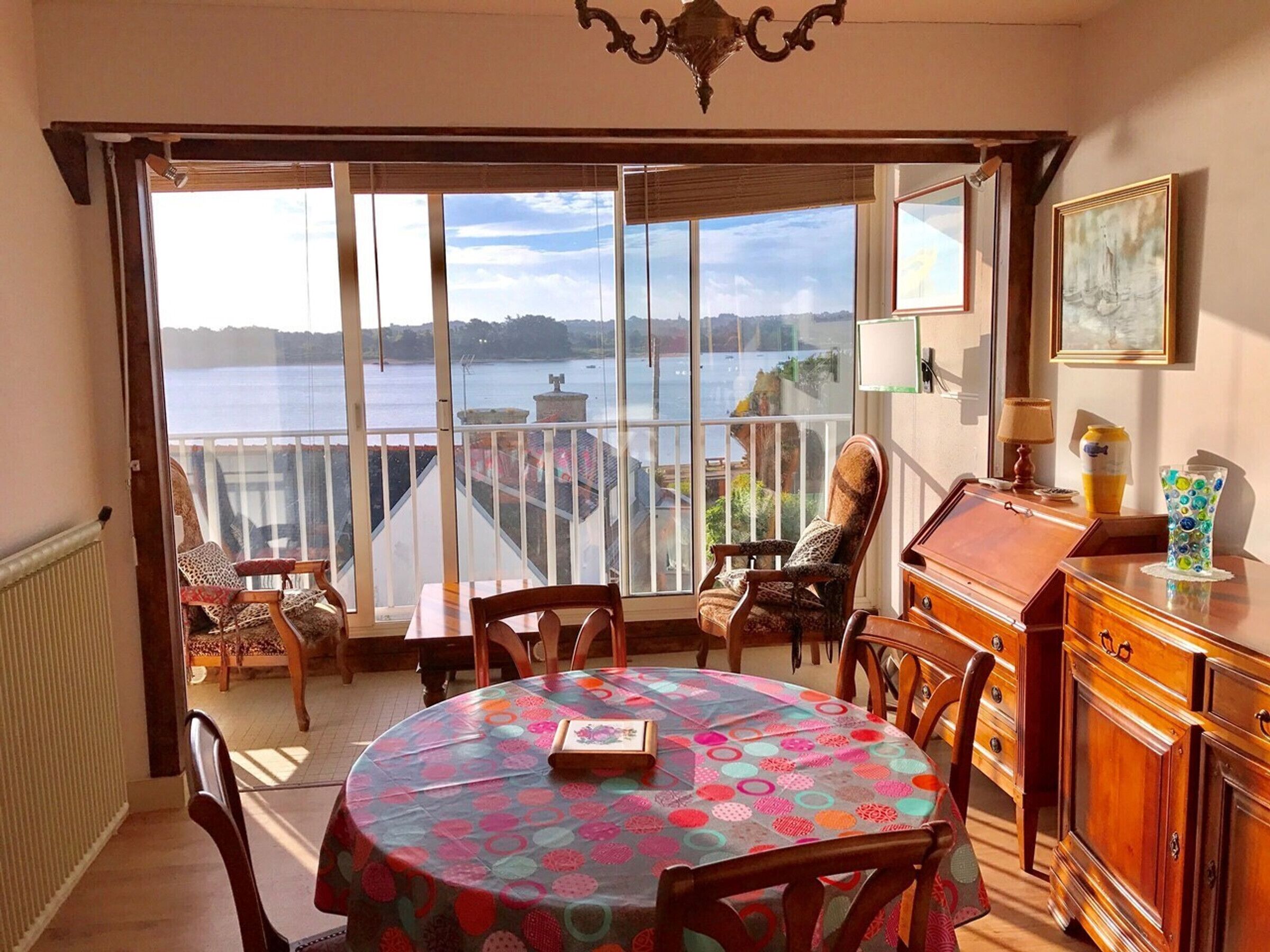 Comedor con vistas a un cuerpo de agua, una mesa redonda con mantel colorido y mobiliario de madera.