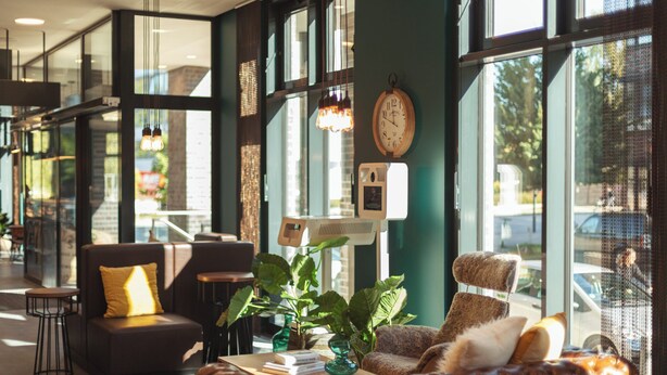 A modern interior with a leather sofa, a small table with books, and a large window with a view of the street. - Holiday Inn - the niu, Bricks Hamburg by IHG (Hamburg)