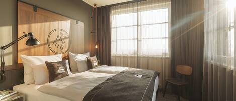 Habitación de hotel con una cama grande, un escritorio con lámpara, una silla y una ventana con cortinas.