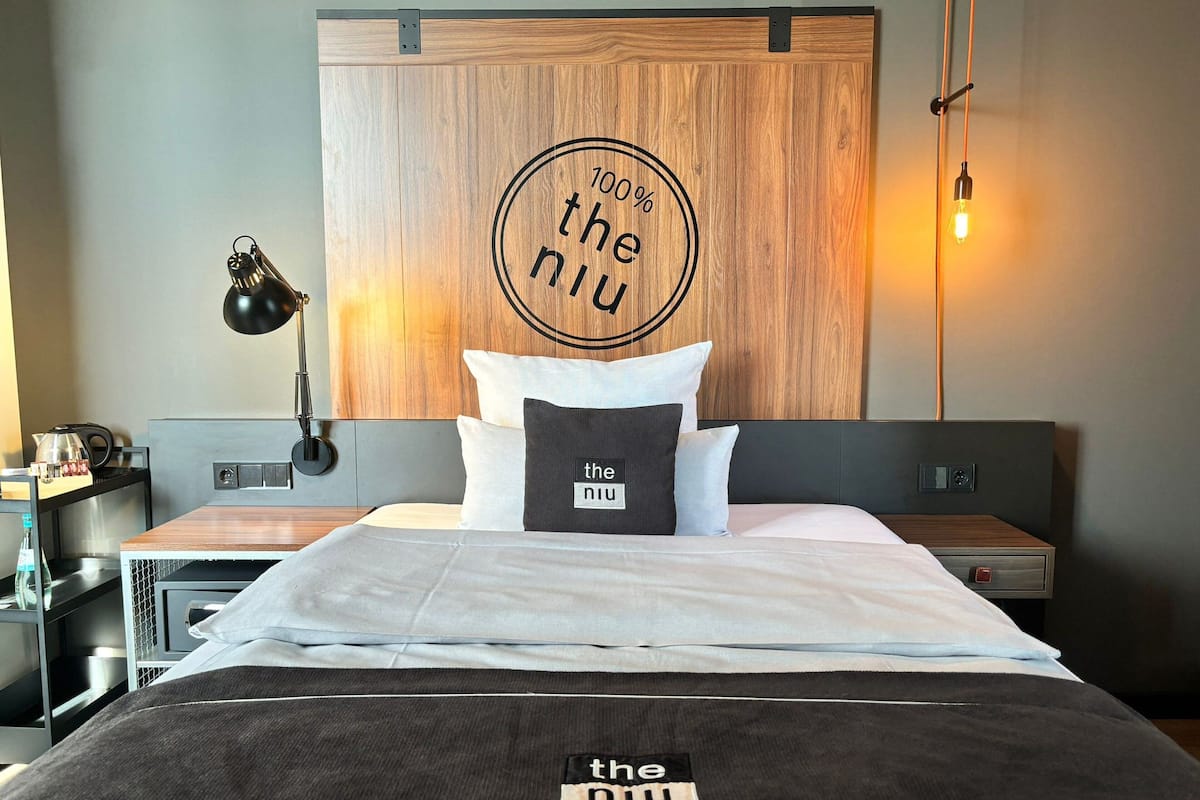 A neatly made bed with a pillow labeled 'the niu,' a bedside lamp, a wooden headboard, and a wall-mounted light fixture.
