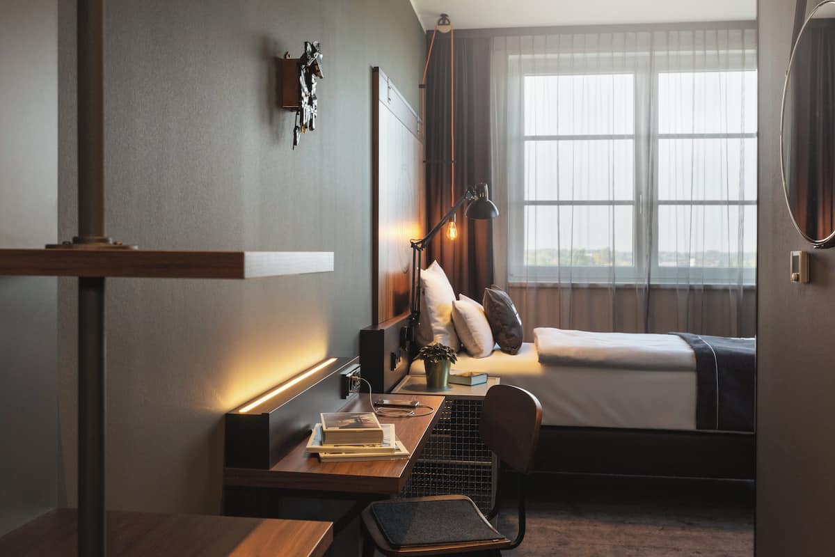 A hotel room with a bed, a desk with a chair, a lamp, and a window with curtains.