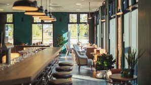 A modern bar area with a long counter, multiple seating options including chairs and a sofa, and large windows providing natural light. - Holiday Inn - the niu, Bricks Hamburg by IHG (Hamburg)