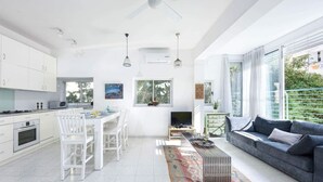 A modern kitchen with white cabinetry, a dining area with white chairs, a grey sofa, and a TV. - Luminous & Cozy 2BR — Your Stylish Urban Retreat (Tel Aviv-Yafo)