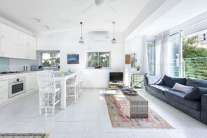 A modern kitchen with white cabinetry, a dining area with white chairs, a grey sofa, and a TV.