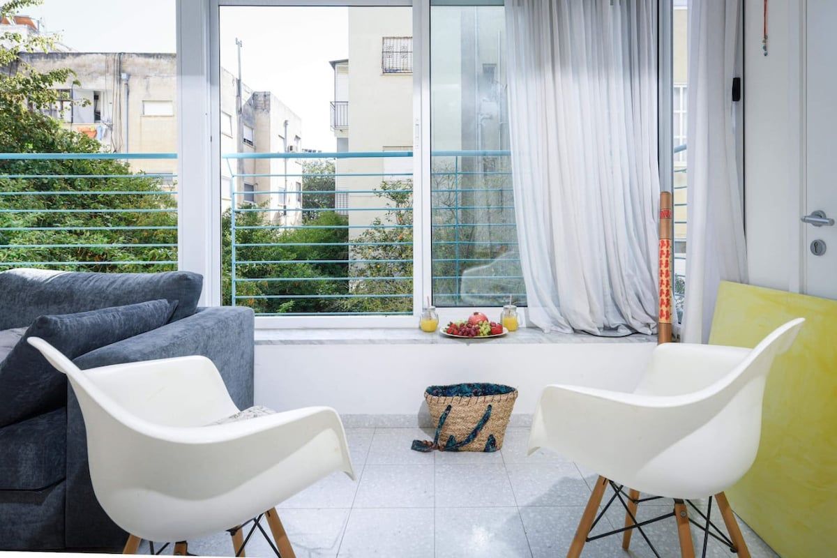 Ein Balkon mit einer grauen Couch, weißen Stühlen, einem gewebten Korb und einer Obstschale.