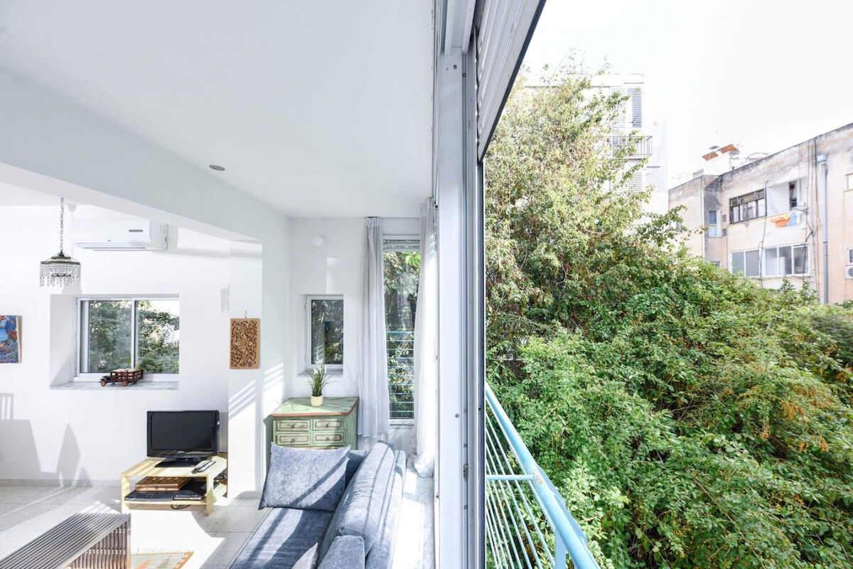 A modern living room with a sofa, television, and a small table. There is a balcony with a view of greenery and buildings.