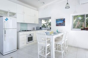 A modern kitchen with white cabinets, a white refrigerator, a dining table with white chairs, and a window with a view of trees.