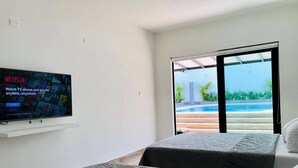 A hotel room with a flat-screen TV displaying the Netflix logo, a bed with a grey patterned bedspread, and a view of a pool and greenery through a glass door. - CARMEN DE APICALA A FANTASY SITE ...! (Anapoima)