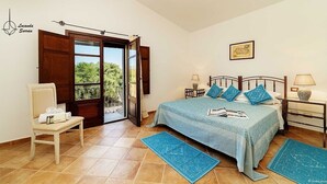 A bedroom with a bed, a chair, a nightstand, a lamp, and a window with a view of greenery. - Locanda In Vigna (Arzachena)