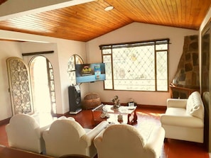 A spacious living room with a wooden ceiling, a television, comfortable seating, and large windows.