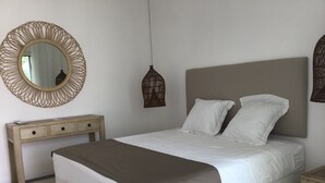 Un lit bien fait, recouvert d’un plaid marron, une table de chevet, une petite table en bois, un miroir rond et un luminaire suspendu.