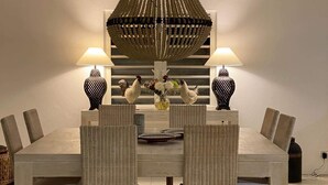 Un espace repas comprenant une grande table rectangulaire, quatre chaises en osier, deux lampes fixées au mur et un luminaire décoratif au plafond.