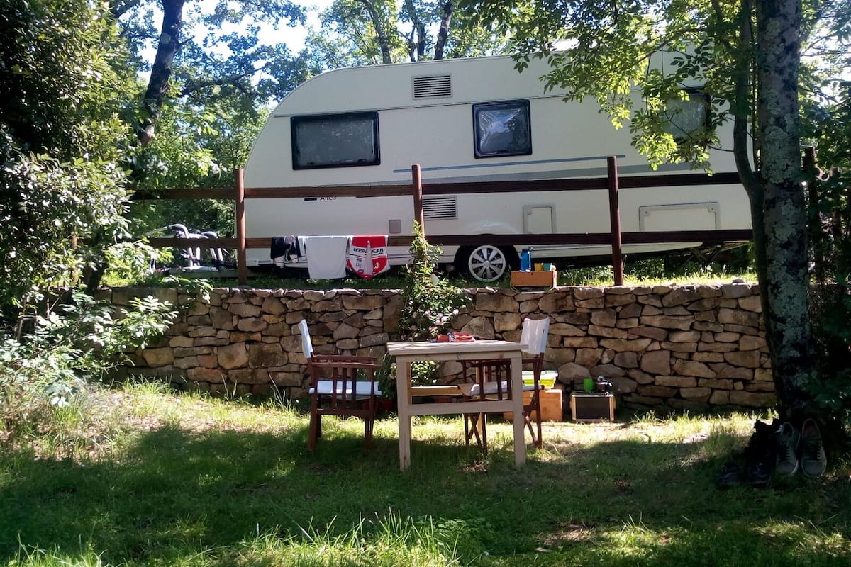Un camping con un muro de piedra, una mesa y sillas de madera, y una caravana estacionada al fondo.