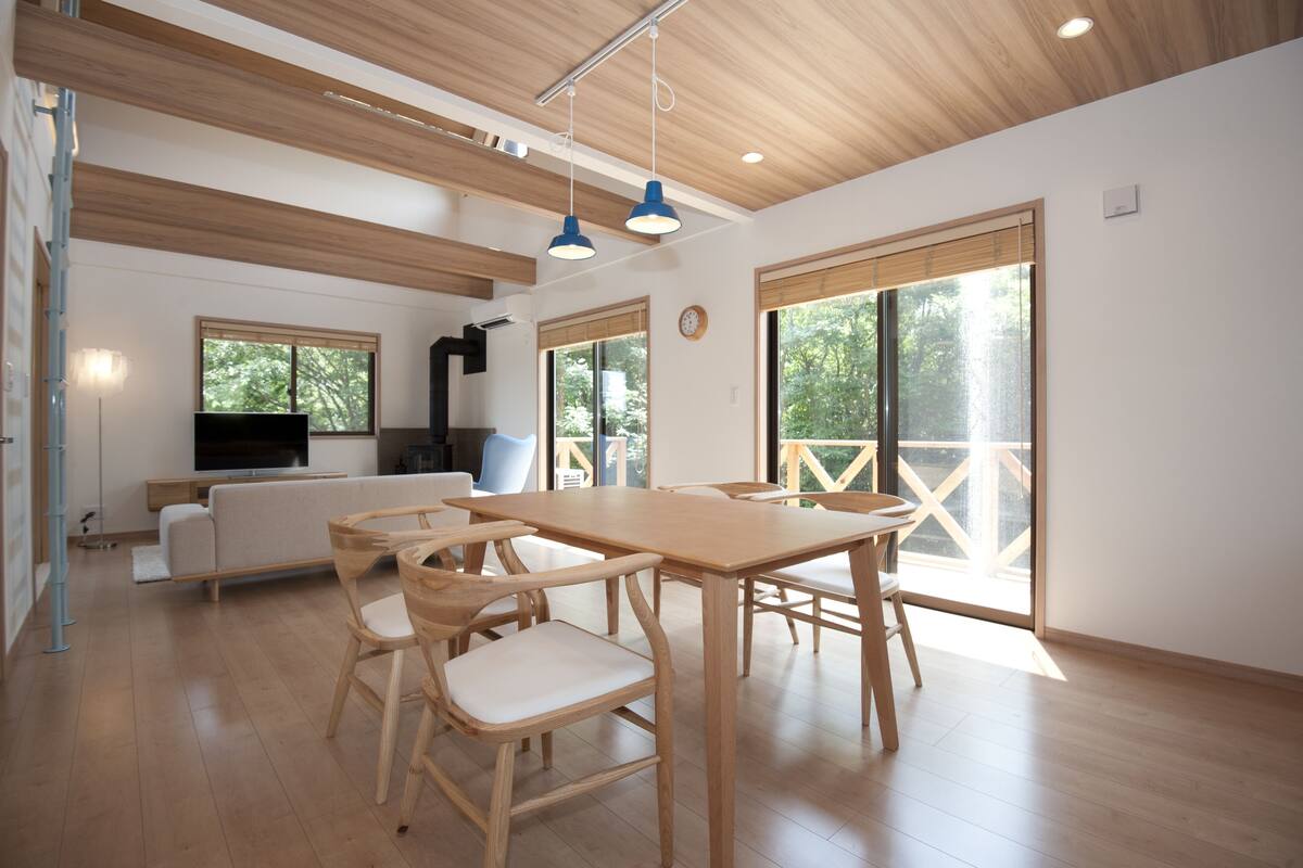 A spacious living area with a wooden dining table, chairs, a sofa, and a TV. Large windows provide natural light and views of greenery.