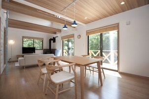 Amplia sala de estar con mesa de comedor de madera, sillas, sofá y televisor. Grandes ventanales ofrecen luz natural y vistas a la vegetación.
