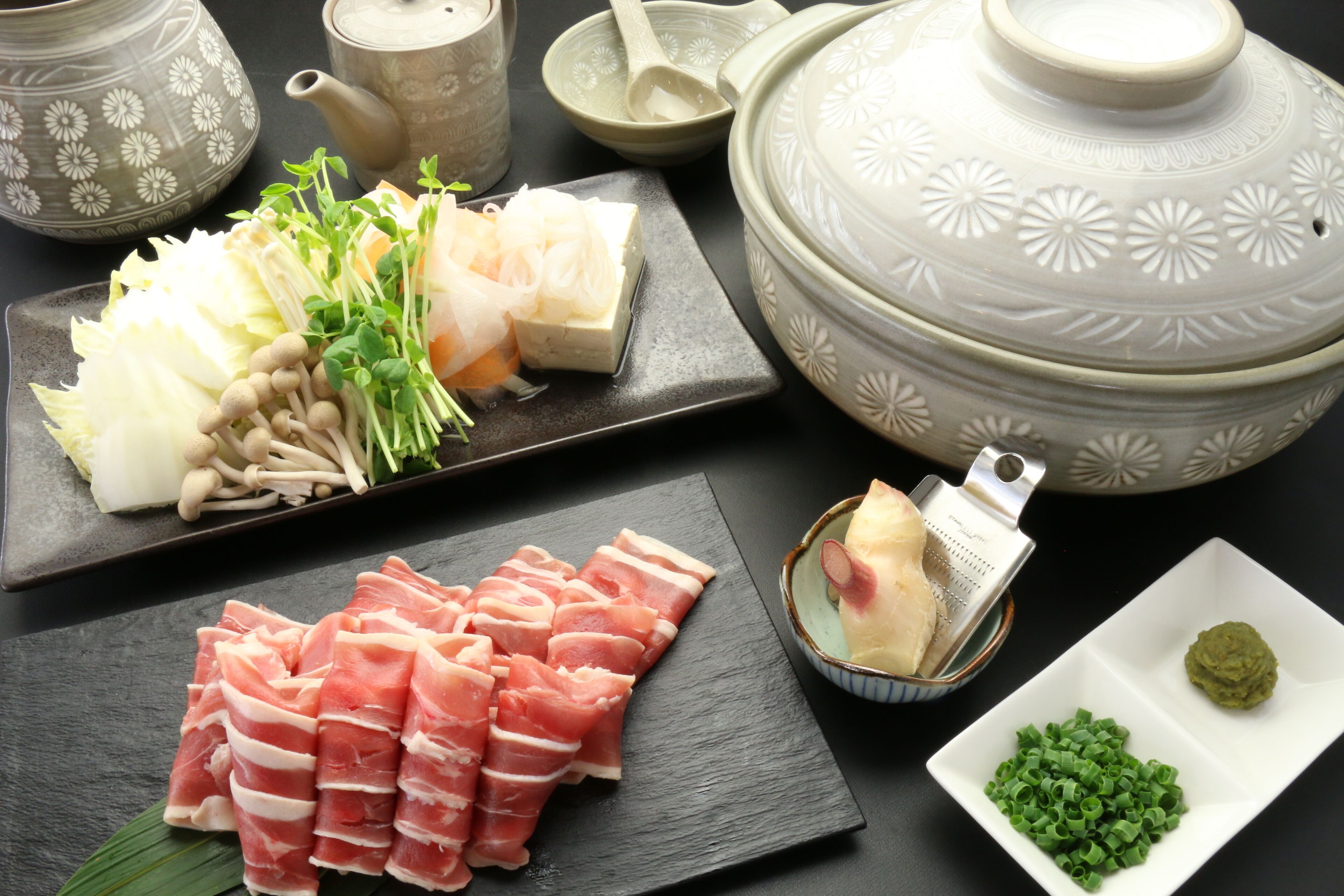 A variety of food items including sliced meat, vegetables, and a bowl of green sauce, arranged on a dark surface with decorative pottery in the background.