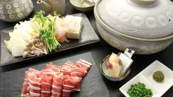 A variety of food items including sliced meat, vegetables, and a bowl of green sauce, arranged on a dark surface with decorative pottery in the background.