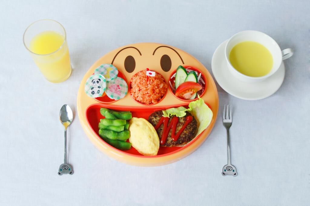 Un plato con un diseño de carita sonriente, que incluye arroz, verduras y una hamburguesa, acompañado de un vaso de zumo de naranja y una taza de té.