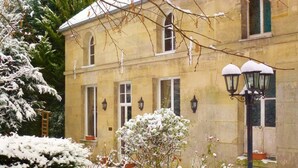 Un bâtiment historique doté d’arcs en plein cintre et d’une façade symétrique, entouré d’arbres couverts de neige et d’un jardin bien entretenu.
