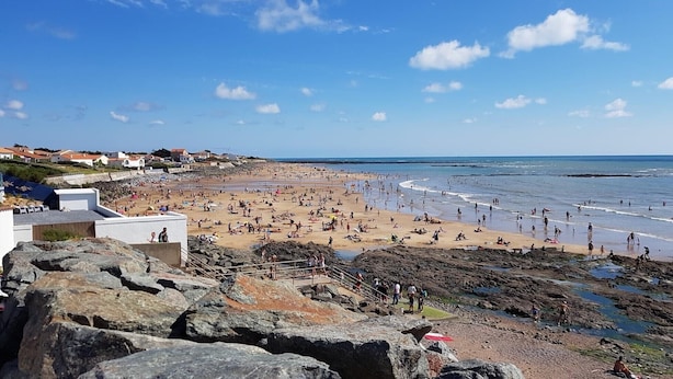 Une plage avec un premier plan rocheux, une zone de plage sablonneuse et des maisons en arrière-plan.