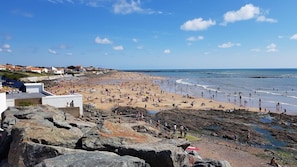 Ein Strand mit felsigem Vordergrund, einem Sandstrand und Häusern im Hintergrund.