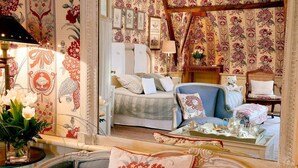Un dormitorio de estilo vintage con papel tapiz floral, una cama con muchos cojines, un sillón azul y una mesita de noche con adornos.