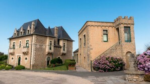 Edificio histórico de piedra con techo empinado y múltiples chimeneas, que presenta una entrada destacada con escalinata de piedra y un pequeño jardín con flores rosadas.