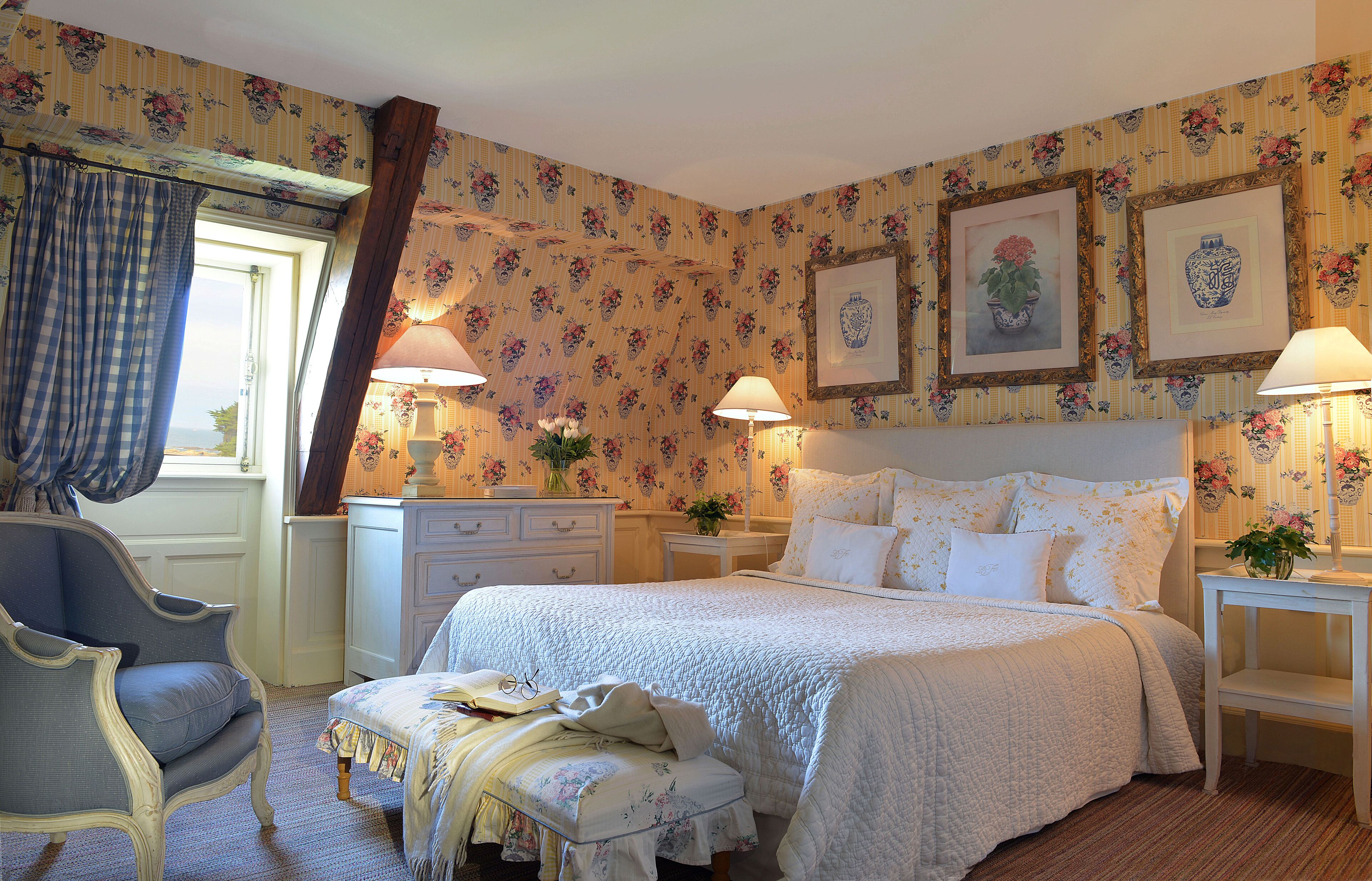 Une chambre à coucher avec un grand lit, deux tables de chevet avec des lampes, une chaise, une commode et un papier peint à motifs floraux.