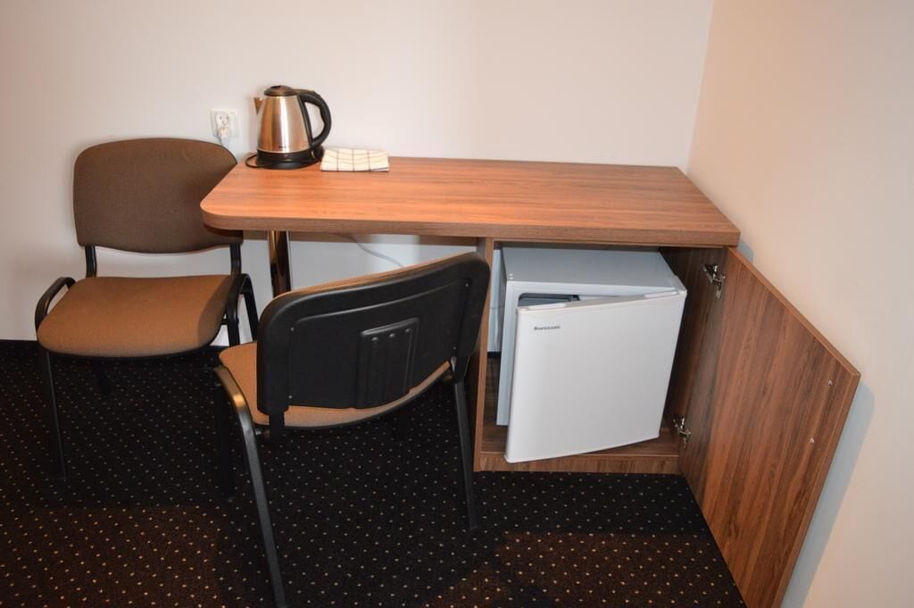 A compact room with a wooden desk, a chair, a small refrigerator, and a kettle.