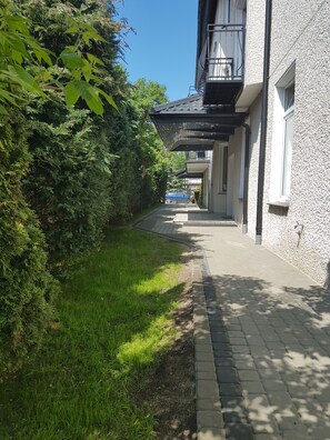 Ein Weg führt zu einem Gebäude mit Balkon, im Hintergrund parkt ein blaues Auto.