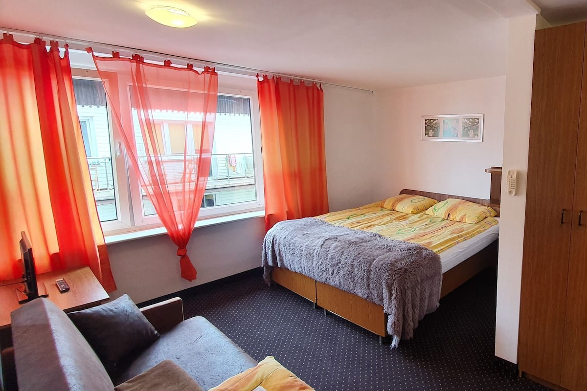 A bedroom with a bed, a wooden wardrobe, a desk with a chair, and a window with curtains.