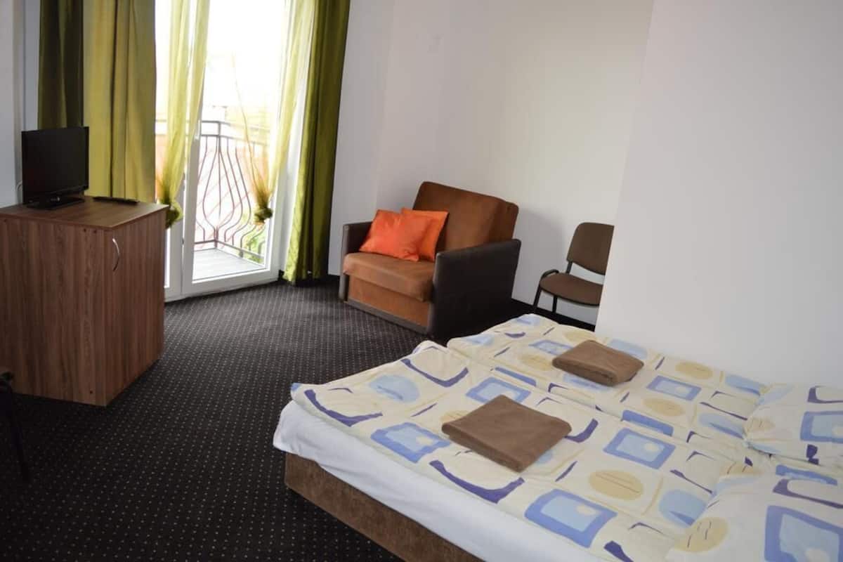 A hotel room with a bed, two chairs, a TV on a wooden cabinet, and a balcony with green curtains.