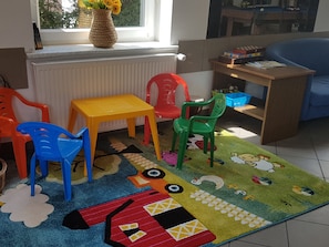 Ein Kinderzimmer mit Fenster, Heizkörper, bunten Teppich und verschiedenen Spielsachen und Möbeln.