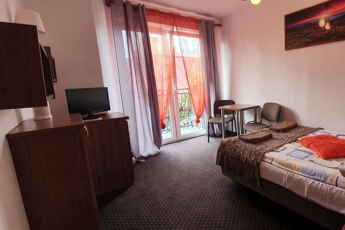 A hotel room with a bed, a desk, a chair, a television, and a balcony with curtains.