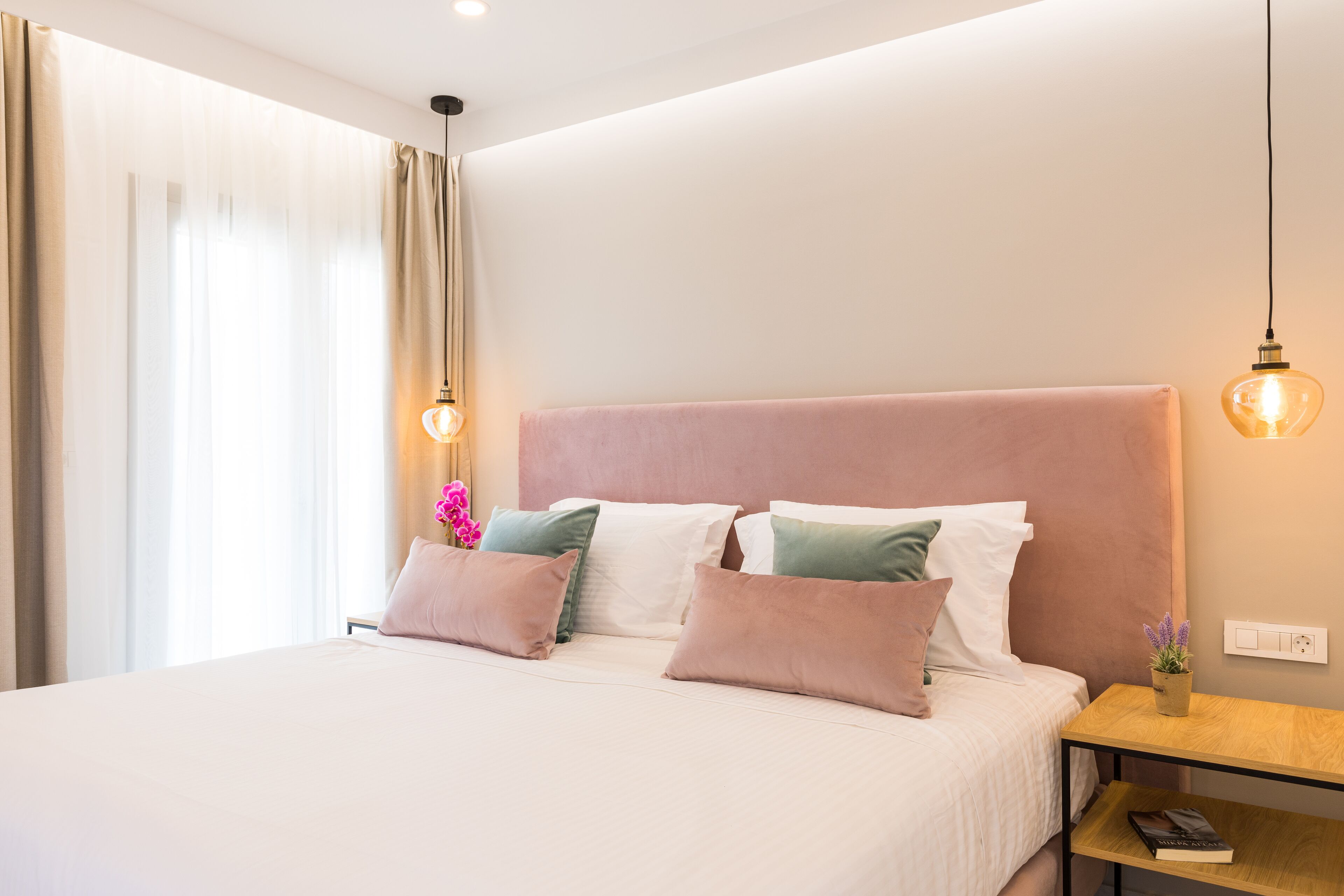 A neatly made bed with a pink headboard, a bedside table with a lamp, and a small potted plant.